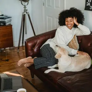 Happy young African American woman sitting on couch and resting with adorable dog with computer and cup of coffee on table in living room